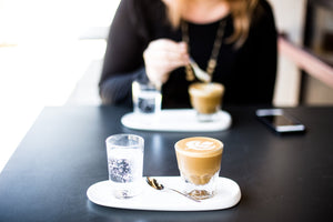 A cortado coffee with latte art served with carbonated water, a woman in the background.