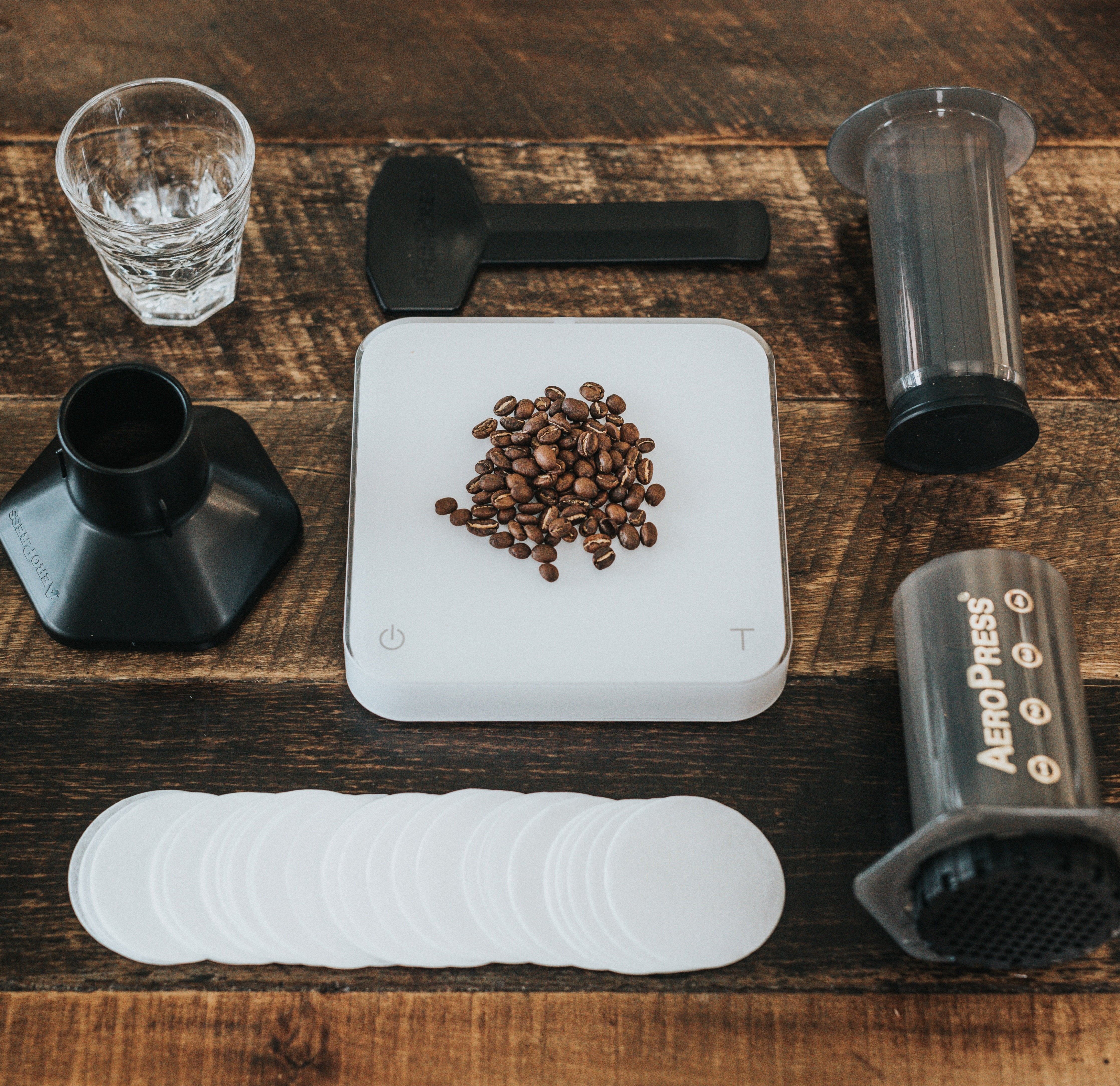 A portable Aeropress set, a glass of water, and Acaia scale with some coffee beans on top.